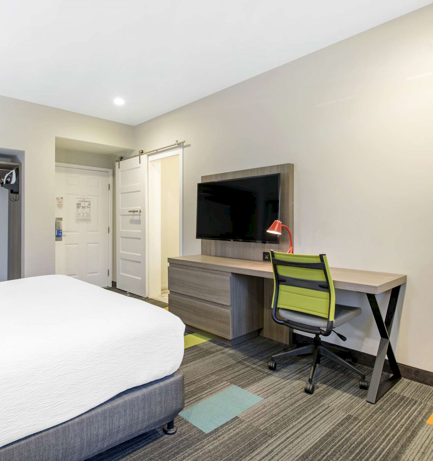 The image shows a modern hotel room with a bed, a desk and chair, a wall-mounted TV, an open closet area, and a doorway leading to another room.