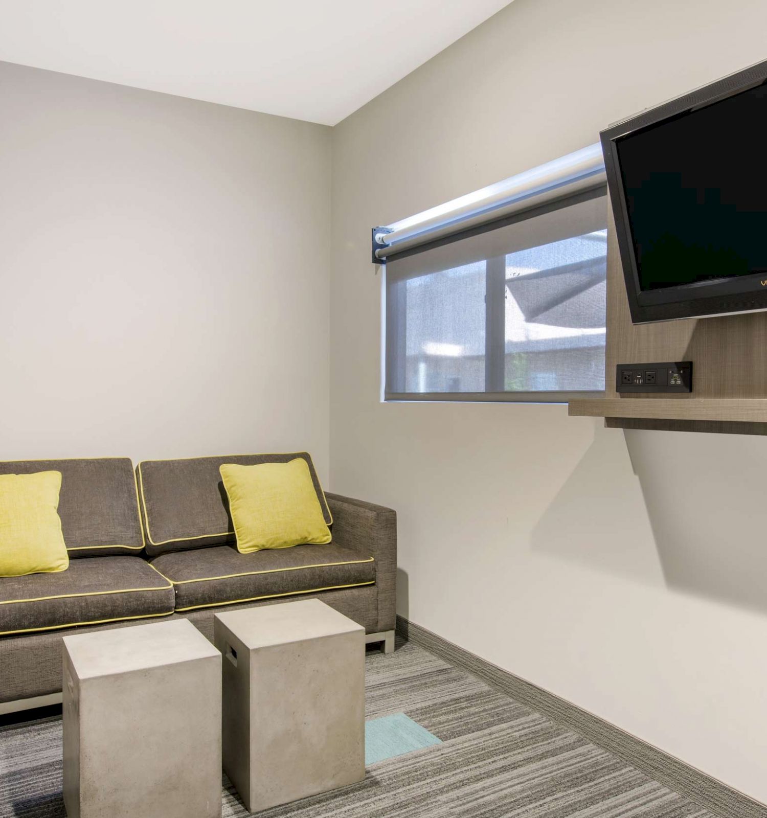 A small room with a gray sofa and yellow pillows, two square tables, a wall-mounted TV, and a window with a roller shade, ending the sentence.