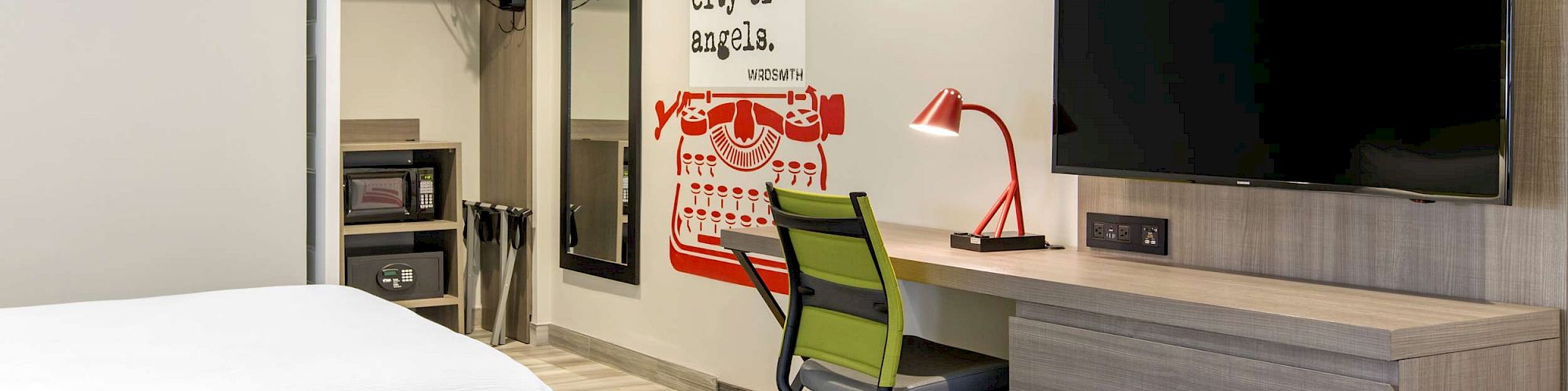 A modern hotel room with two beds, a wall-mounted TV, a desk with a red lamp, and a typewriter-themed wall art with text.