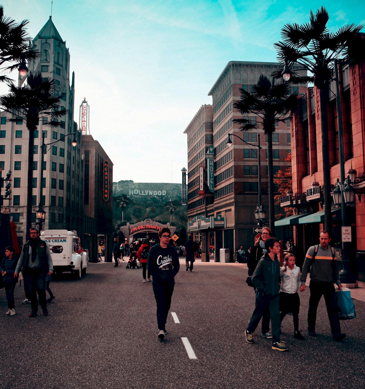 The image shows a busy street scene with people walking, surrounded by tall buildings and palm trees, and a distant sign resembling 