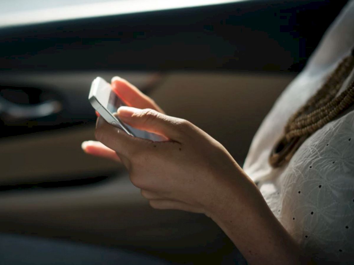 A person is using a smartphone, sitting by a car window, wearing a light-colored top and a beaded necklace, with natural light coming through.