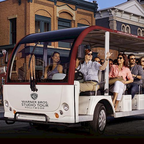 A group of people are seated in an open-air tram with "Warner Bros. Studio Tour" branding, riding through a studio lot with building sets in the background.