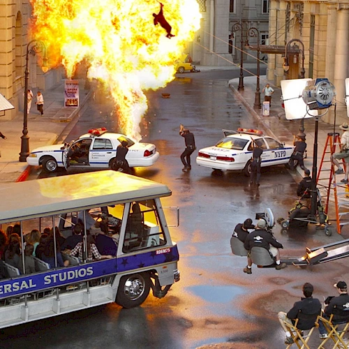 The image shows a film set with a tram from Universal Studios, an explosion, police cars, crew members, and equipment being used for shooting scenes.