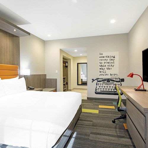 The image shows a modern hotel room with a large bed, desk, flat-screen TV, and a wall quote featuring a typewriter.