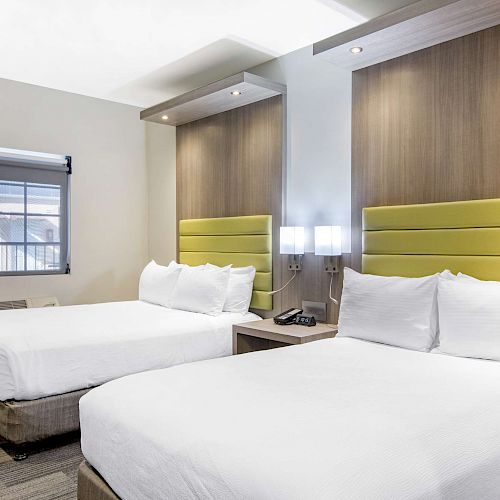 The image shows a modern hotel room with two neatly made beds, green headboards, bedside lamps, and a window with a view.