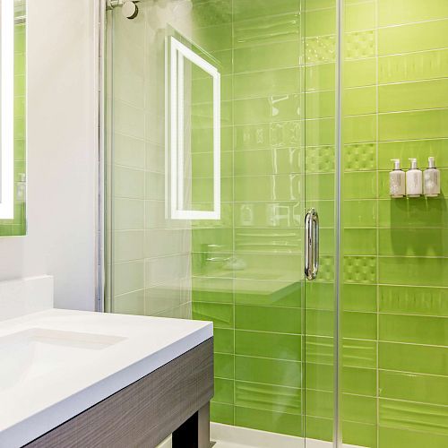 A modern bathroom with a green-tiled shower, glass doors, a white sink, illuminated mirror, and wall-mounted soap dispensers.