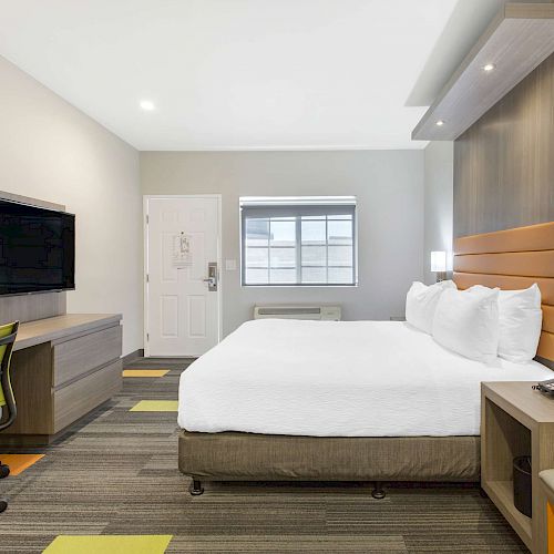 A modern hotel room with a large bed, TV, desk, chair, and nightstand. The room appears bright and neatly organized.