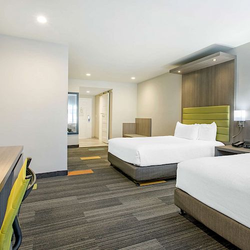 A modern hotel room with two beds, a wall-mounted TV, a desk with a chair, and contemporary lighting fixtures.