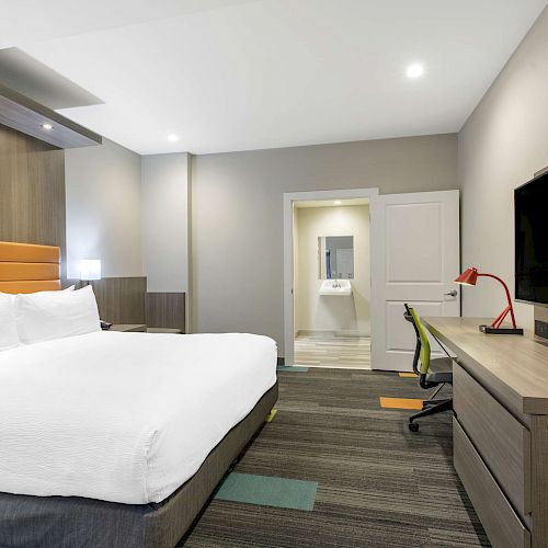 A modern hotel room with a large bed, wall-mounted TV, desk, chair, and minimalist decor in neutral tones.