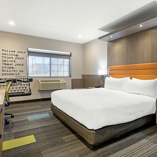 A modern hotel room with a king-sized bed, desk, TV, window, and motivational wall art featuring a typewriter.