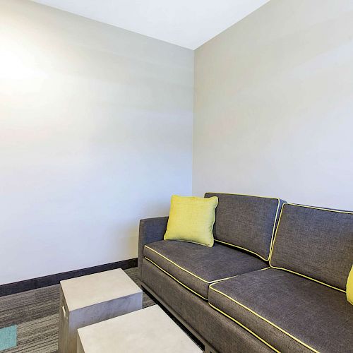 The image shows a modern living area with a grey couch accented by yellow pillows, a wall-mounted TV, and minimalist decor.