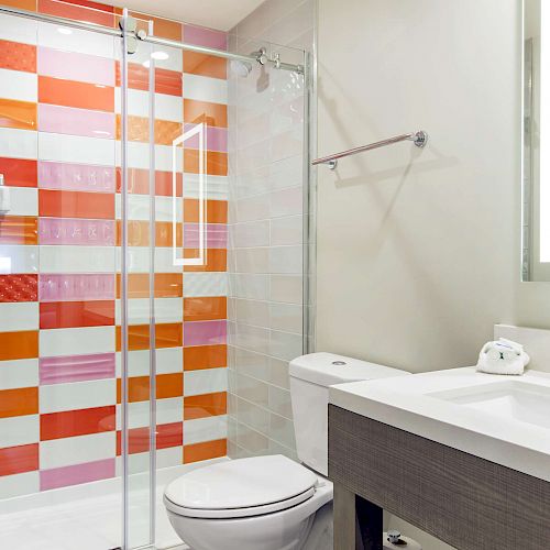 A modern bathroom with a colorful tiled shower, glass doors, a white toilet, and a vanity with a sink, faucet, and lighted mirror ends the sentence.