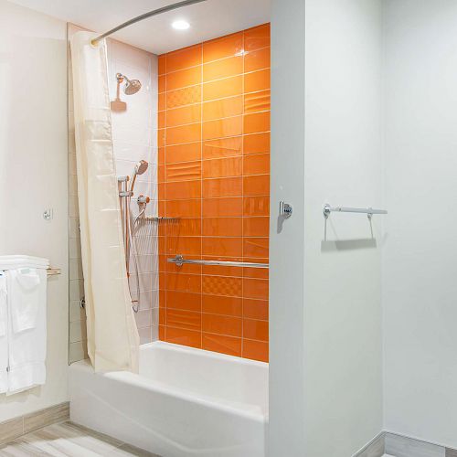 A modern bathroom with a bathtub and shower area featuring orange tiles, a sink with a mirror, and a toilet with towels hanging nearby.