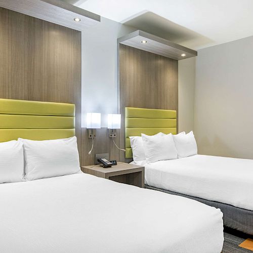 The image shows a modern hotel room with two double beds, white linens, green headboards, a bedside table, and wall-mounted lamps between the beds.