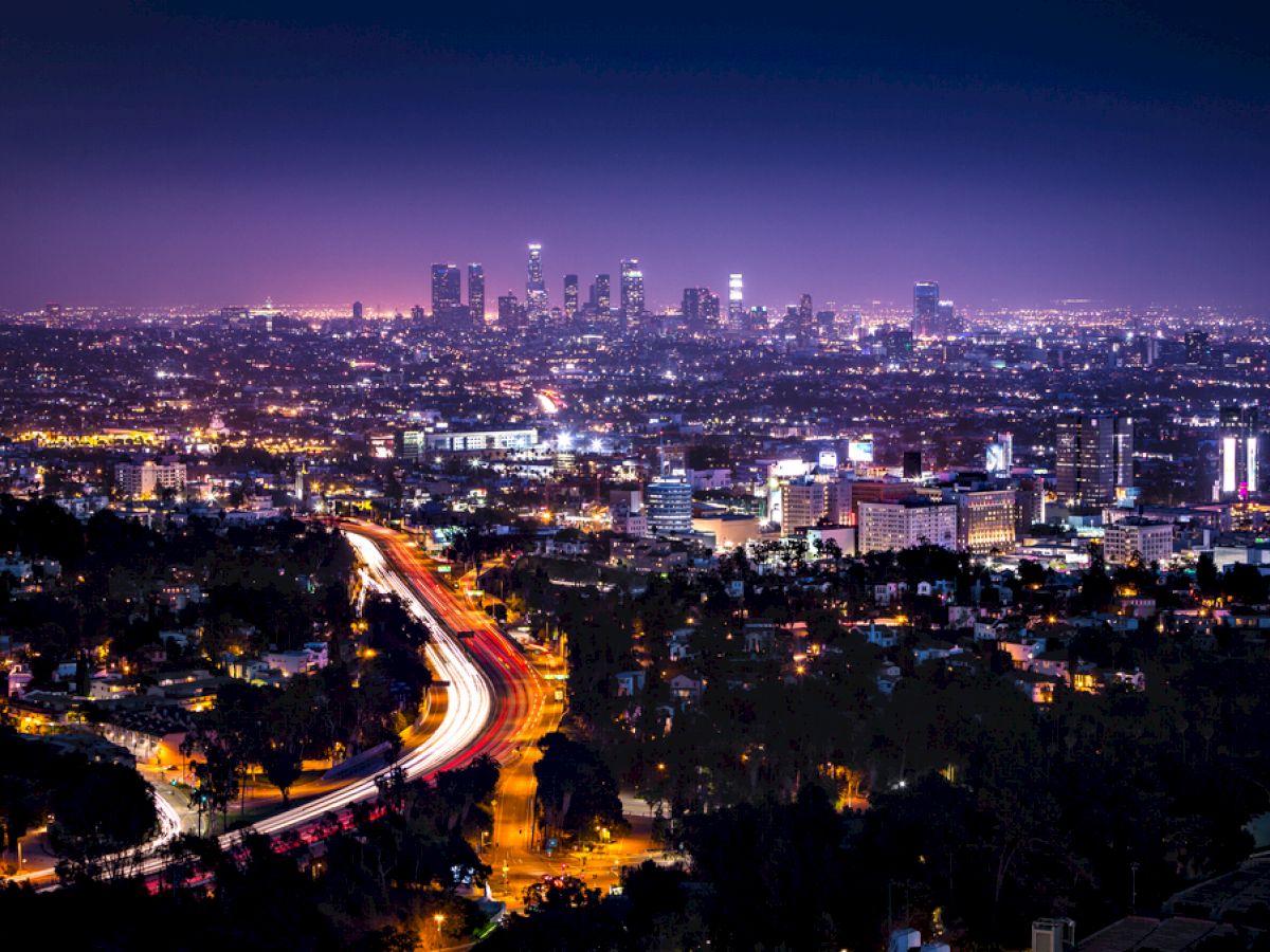 A dazzling night view of a sprawling cityscape with brightly lit skyscrapers and a winding highway illuminated with moving vehicle lights.