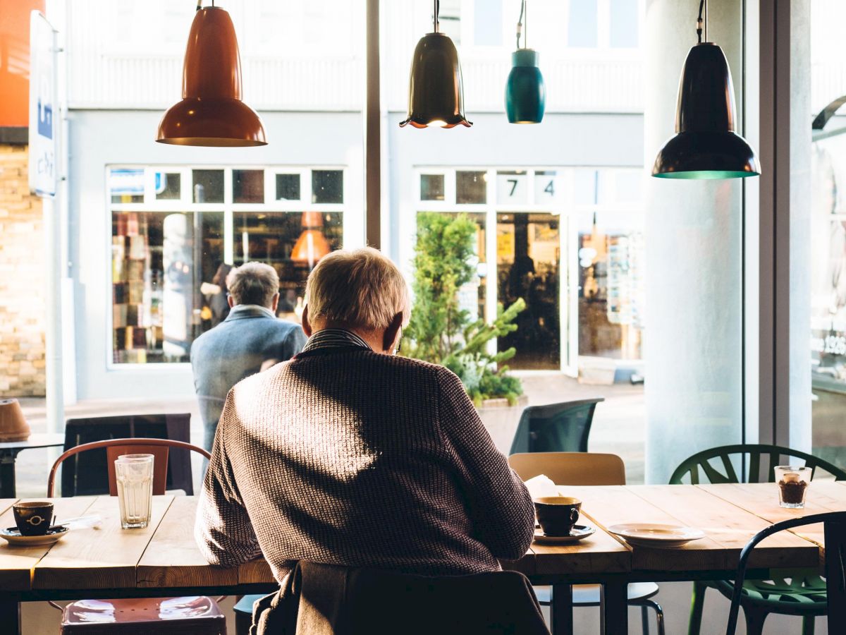 A person sits alone at a café table with coffee, looking out the window; another person is seated outside in the background.