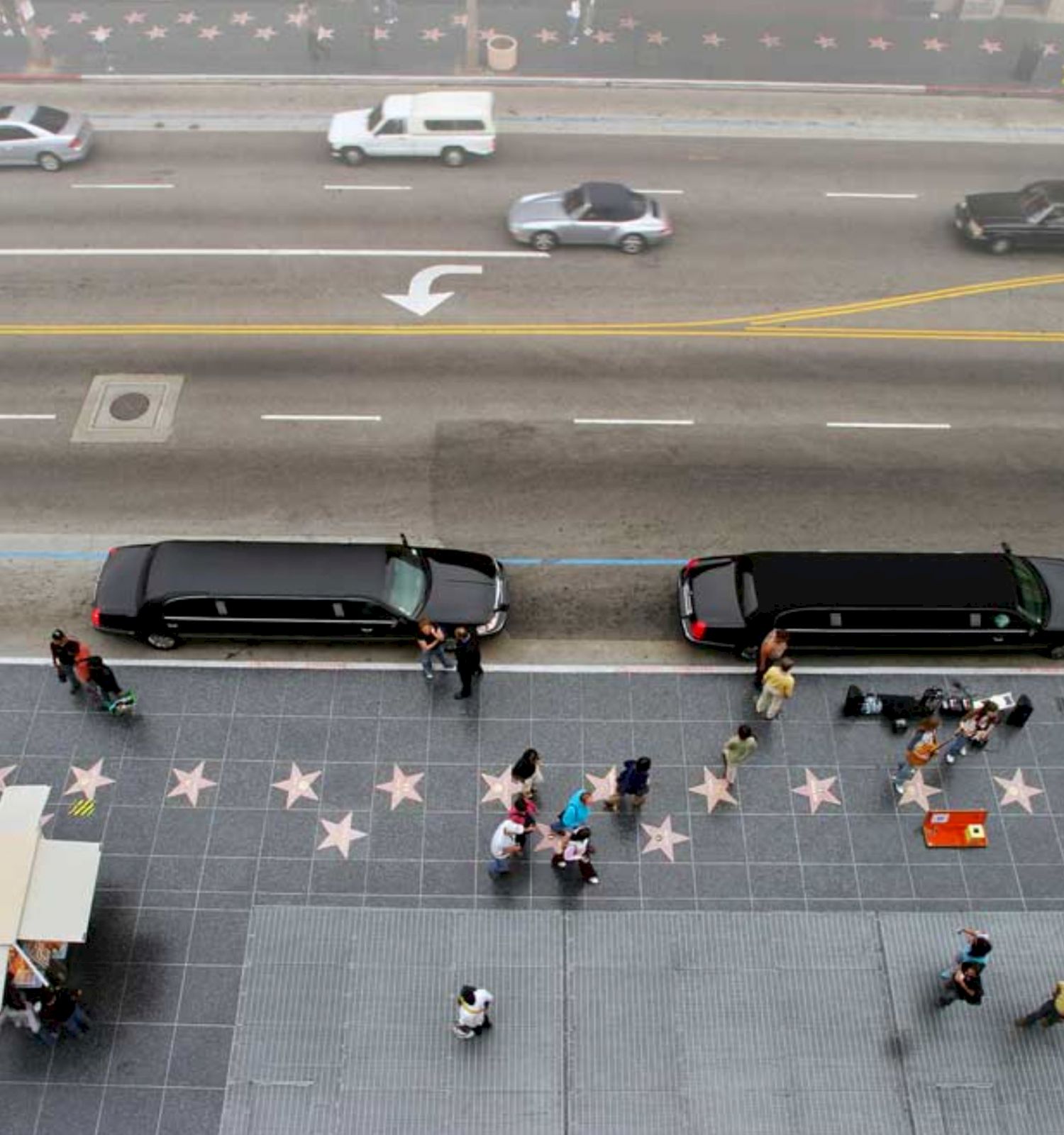 An aerial view of a street with two limousines, pedestrians, and the Hollywood Walk of Fame with star plaques on the sidewalk.