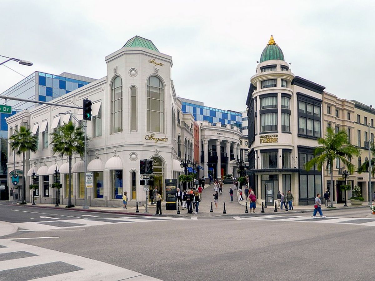 An urban street scene with people, palm trees, and tall, ornate buildings, including a shop and crosswalk with a sign reading 