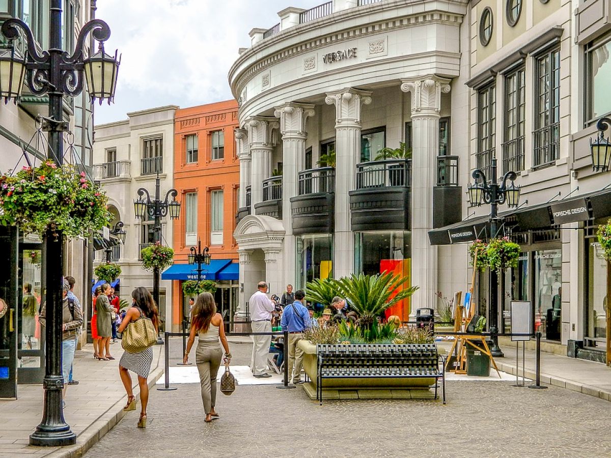 A bustling outdoor shopping area with people walking, surrounded by elegant buildings and hanging flower baskets, featuring classic architecture.