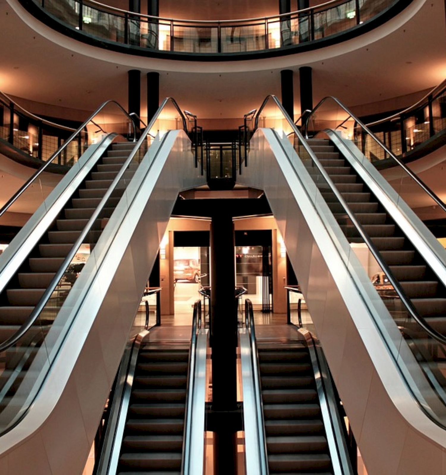 The image depicts multiple escalators inside a modern indoor shopping mall, surrounded by various commercial spaces and lit with ambient lighting.