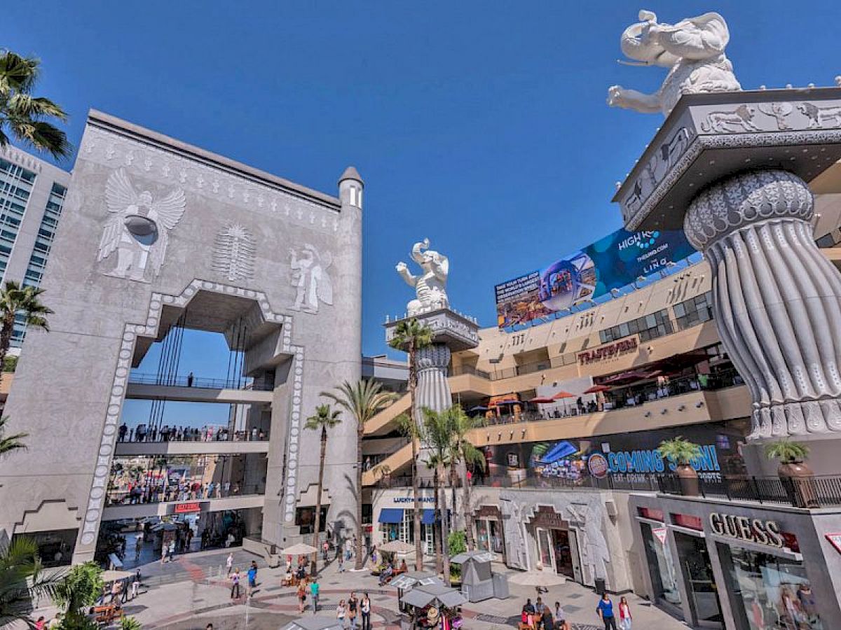 The image shows a plaza with large statues, a retail complex, and people walking around. The architecture has an ancient motif with elephant sculptures.