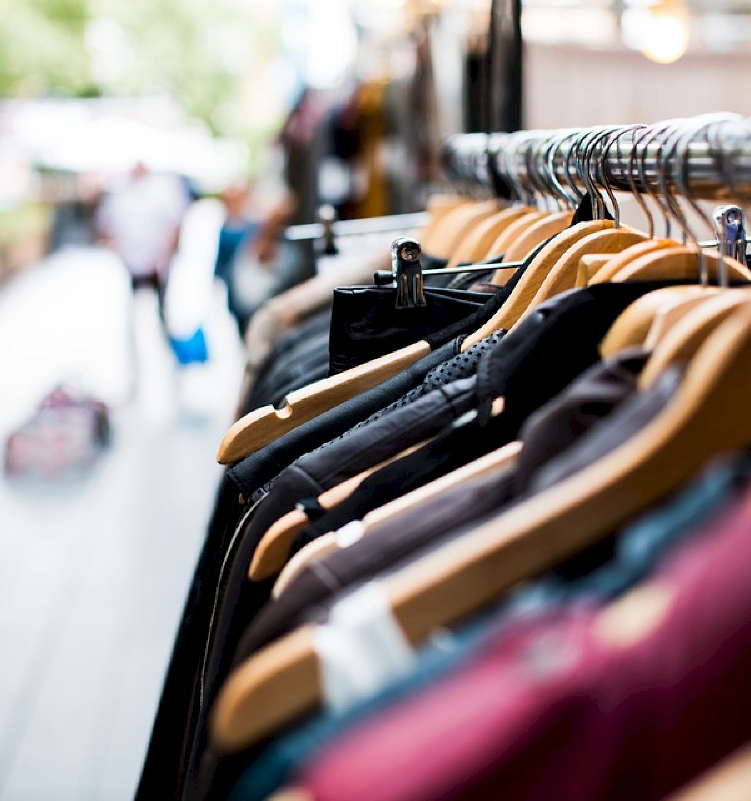 The image shows a row of clothes hanging on wooden hangers on a metal rack, with a blurred background of what appears to be a street scene.