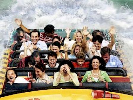 A group of people is on a water ride, splashing down a track with excitement and raised hands. The ride is labeled 