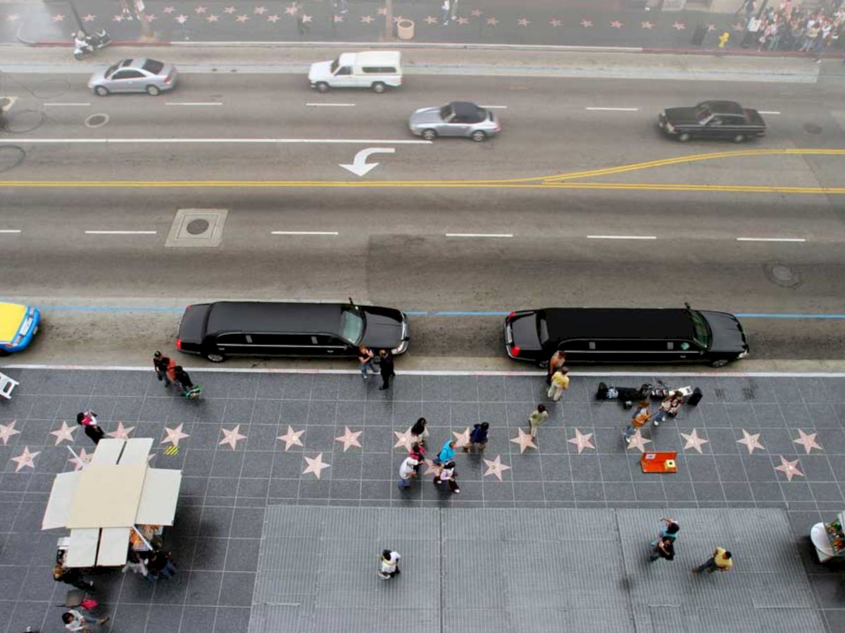 A street view with cars, two black limousines, and people on a star-studded sidewalk, possibly the Hollywood Walk of Fame.