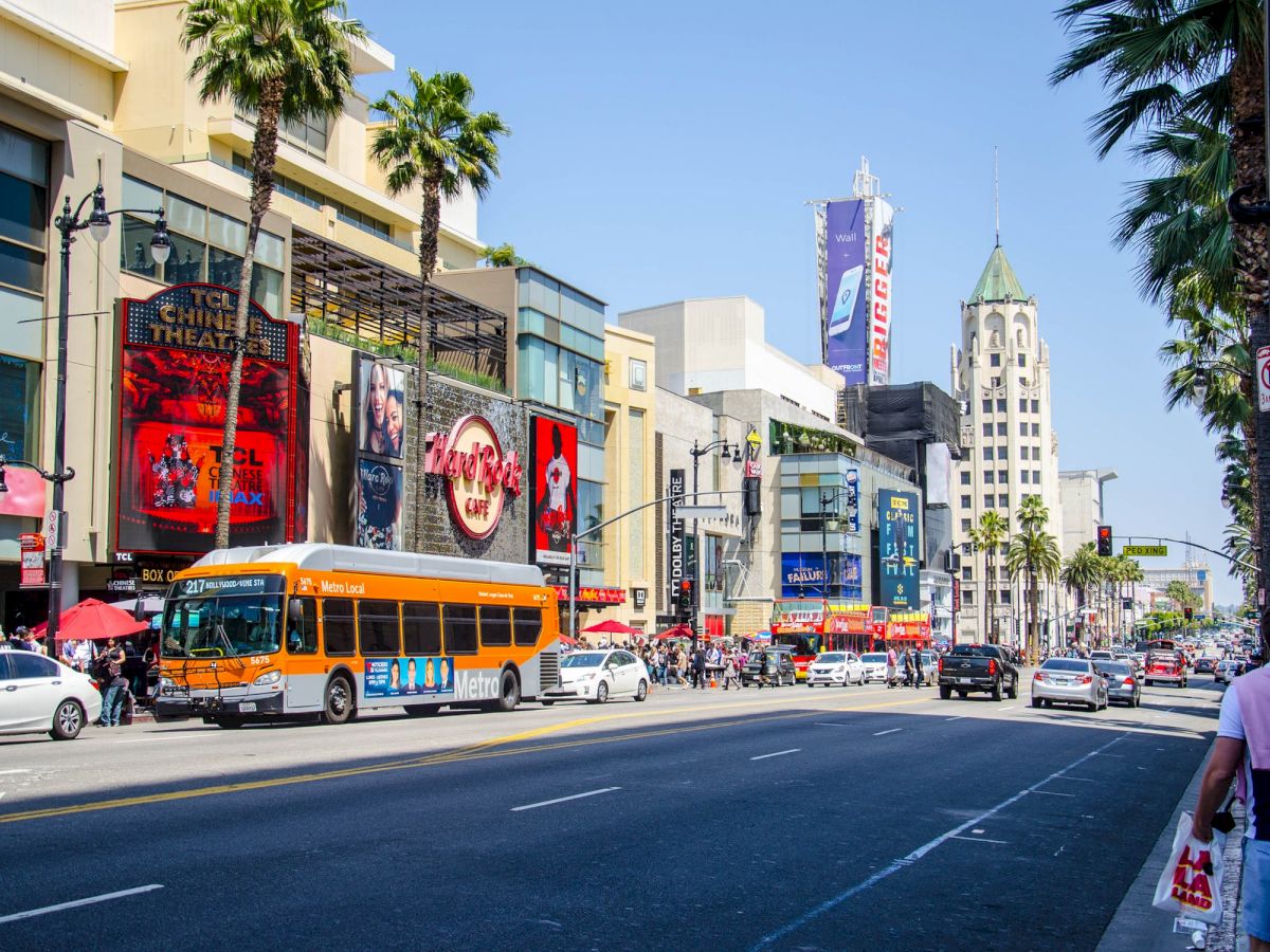 A busy street scene with a bus, people, cars, and shops. Tall buildings and palm trees line the street in bright daylight, ending the sentence.