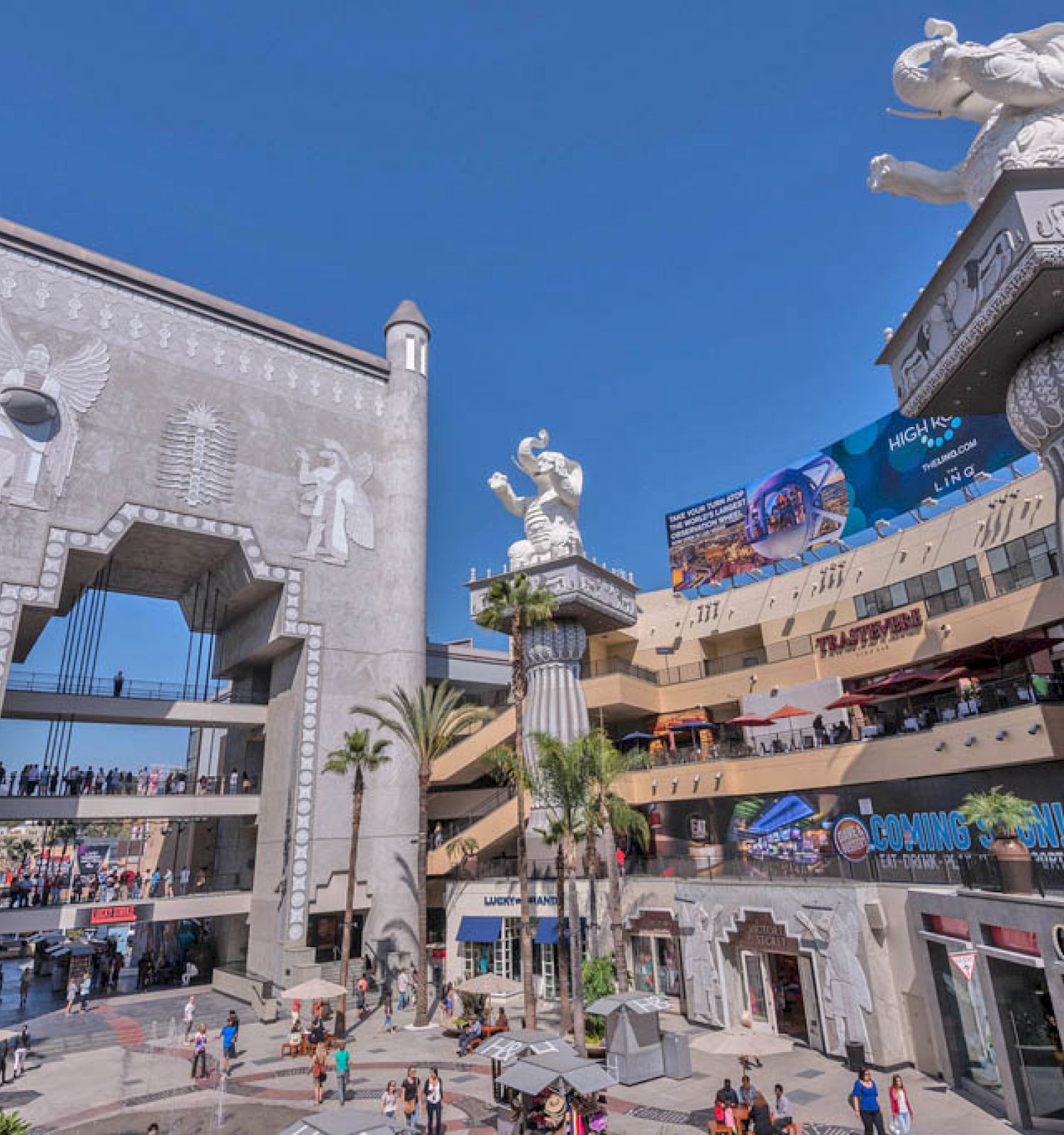 An outdoor shopping and entertainment complex with a grand archway, crowd of people, palm trees, and large elephant sculptures atop columns.