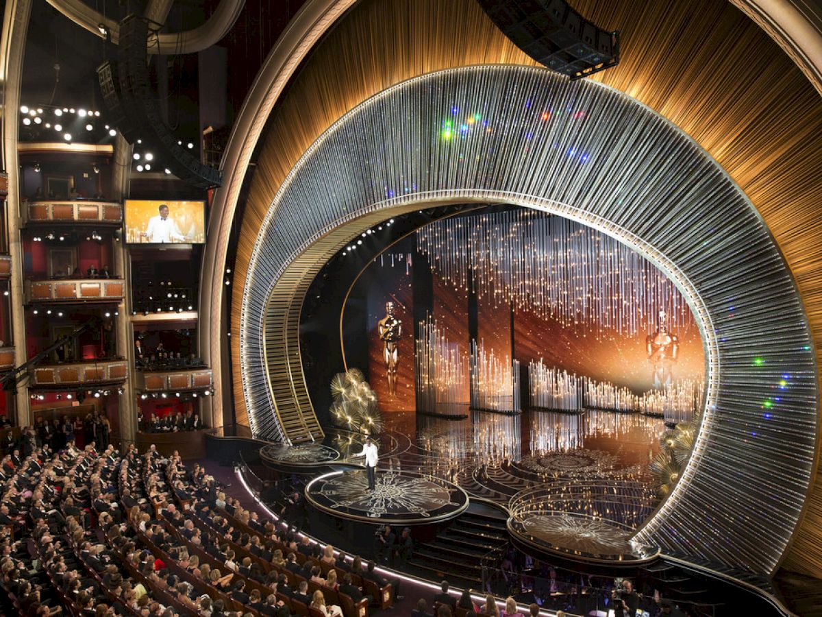 A grand theater scene with a stage, an ornate backdrop, an audience, and a presenter. The setting looks like an awards ceremony.