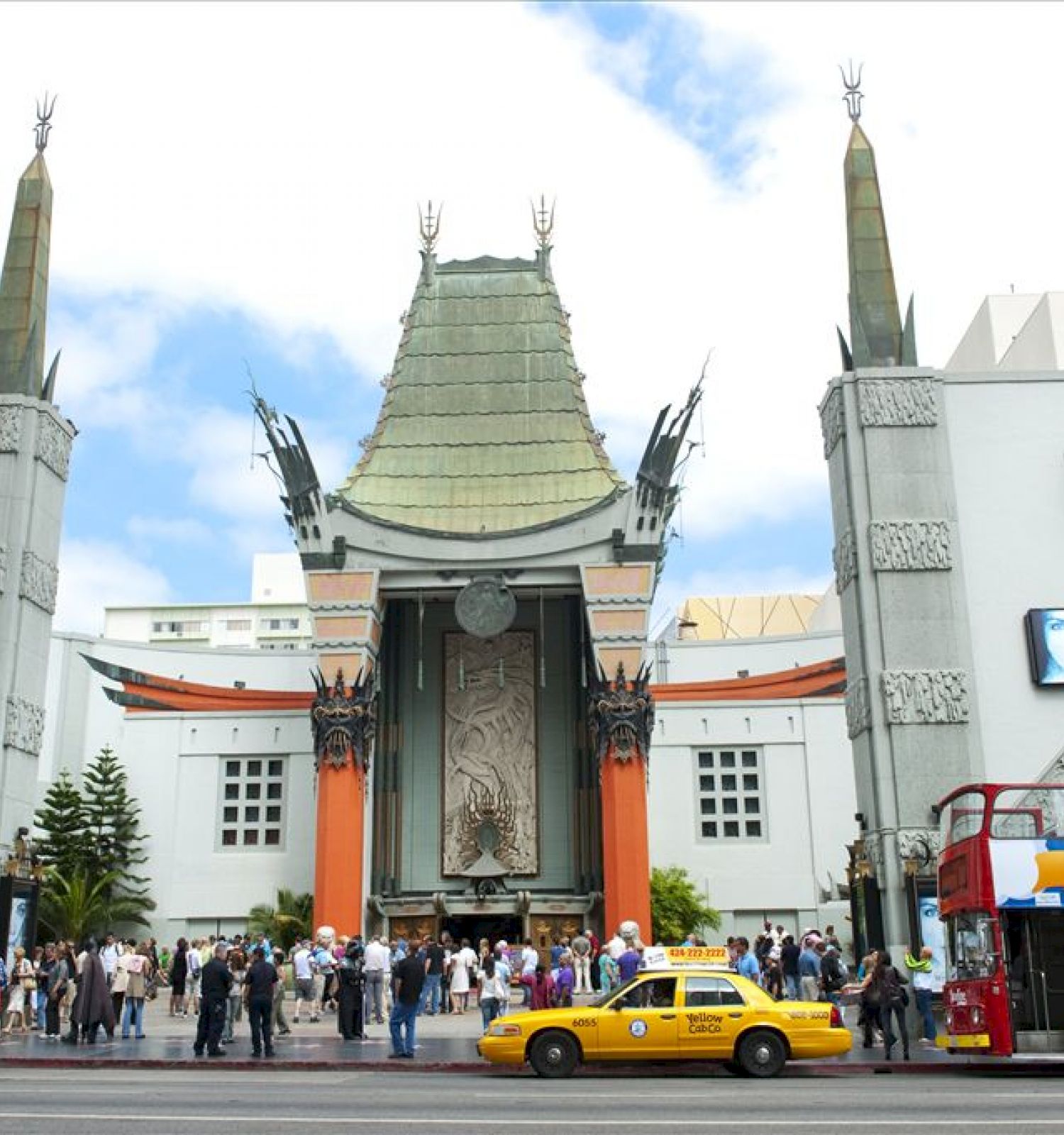 Many people gather outside a historic theater with ornate architecture, some taxis, and a double-decker bus nearby, under a clear sky.