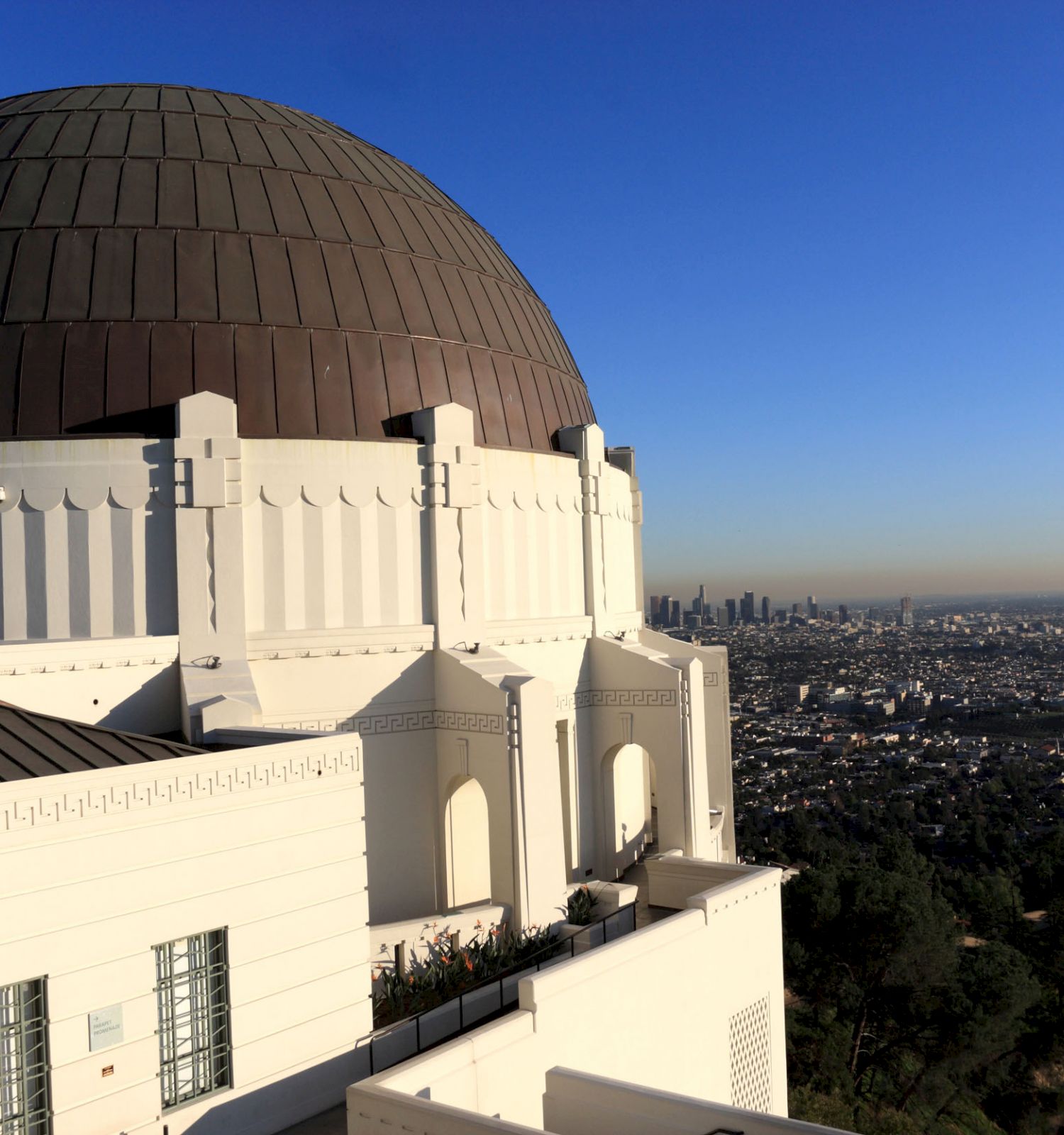 The image shows a white observatory with a large dome, overlooking a sprawling cityscape under a clear blue sky.