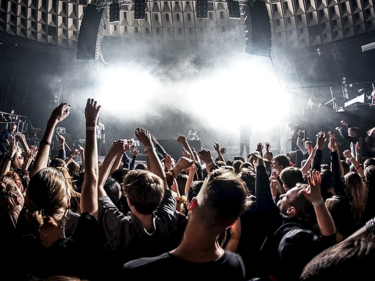 A lively crowd is enjoying a concert with raised hands, surrounded by bright stage lights and a dark, atmospheric venue.