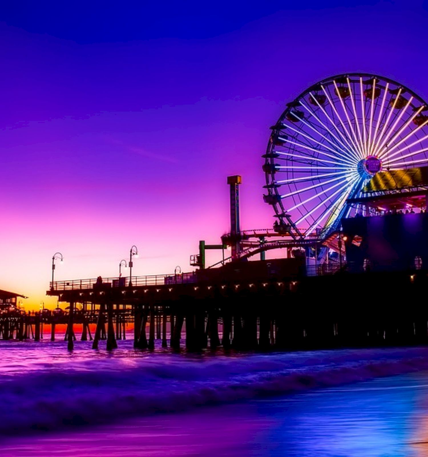 A lit Ferris wheel stands at the end of a pier, surrounded by vibrant colors of a sunset, reflecting off the water's surface.