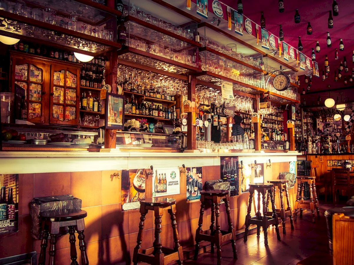 A cozy bar with wooden stools, a well-stocked counter, hanging beer glasses, and decorative bottles on the ceiling. Warm lighting sets a welcoming mood.