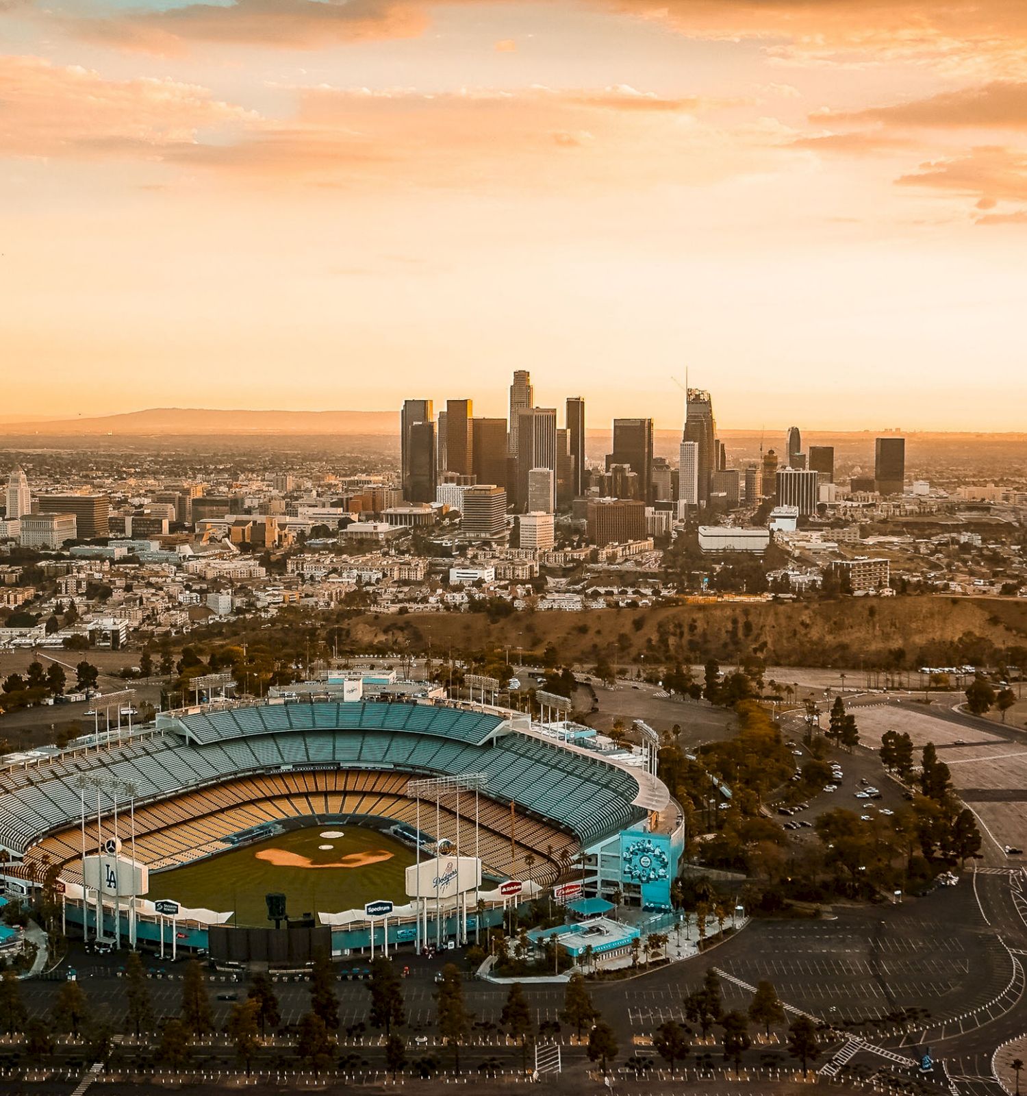 An aerial view of a large baseball stadium with a city skyline in the background, under a beautiful sunset sky.