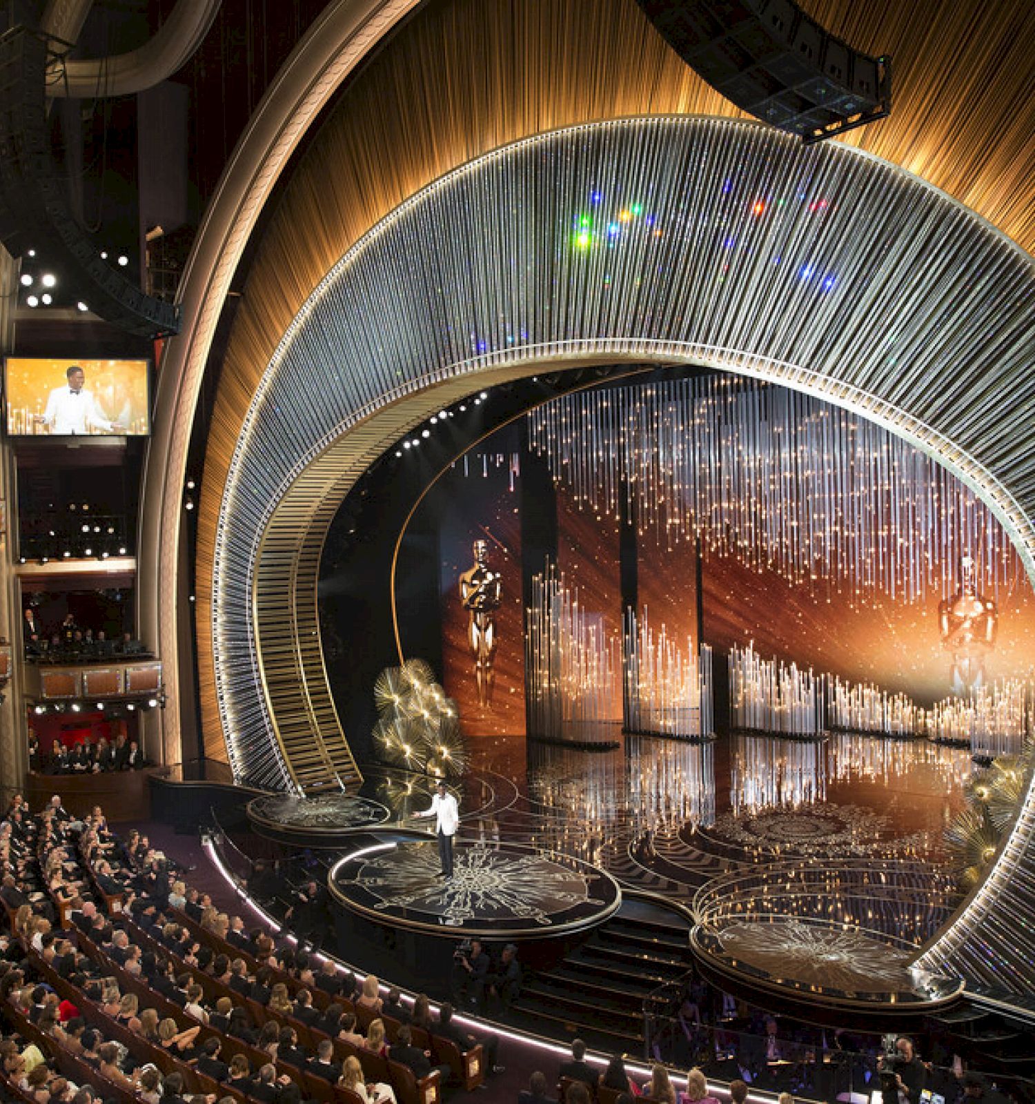 The image depicts a grand awards ceremony in a large theater, with a beautifully decorated stage and an audience seated.