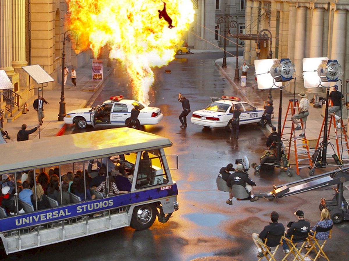 A movie scene being filmed on a city street with an explosion and police cars; tourists observe from a Universal Studios tram.