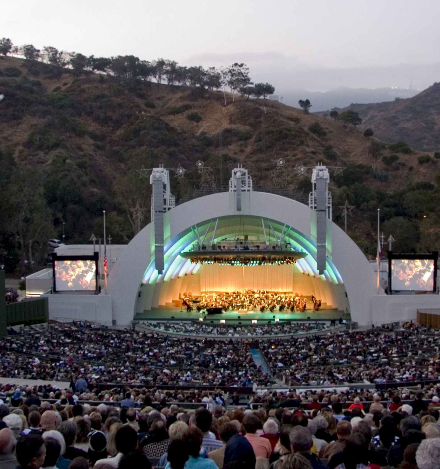 An outdoor concert venue nestled in a hilly landscape with a large audience facing the stage, which is illuminated and flanked by two screens.