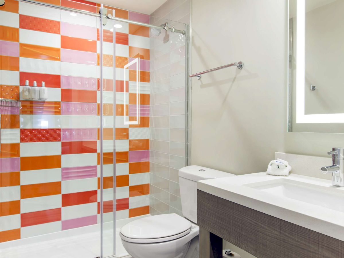 A modern bathroom with white fixtures, an illuminated mirror, and a shower featuring a bold, multicolored tile wall pattern in orange and red hues.