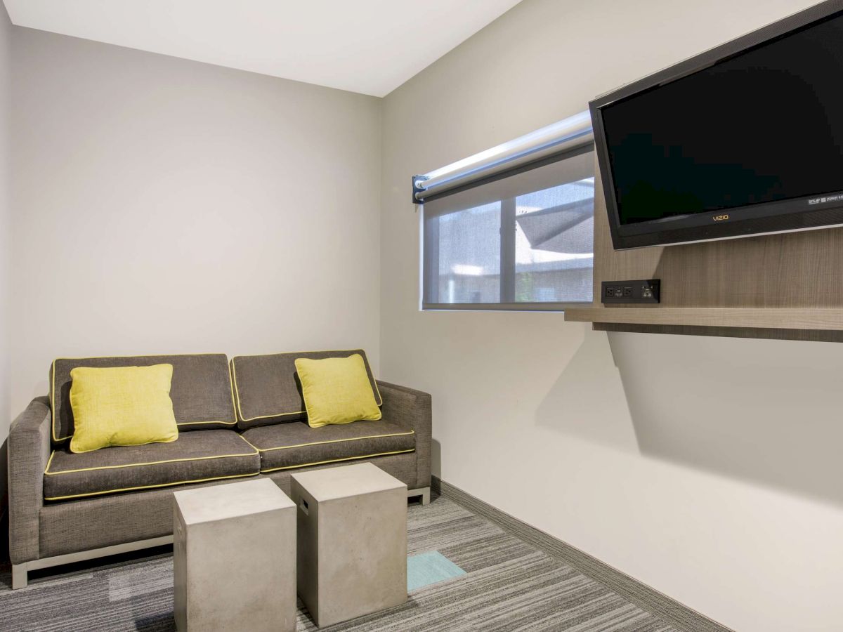 A small room with a sofa, two yellow pillows, two cube-shaped tables, a wall-mounted TV, and a window with a blind.