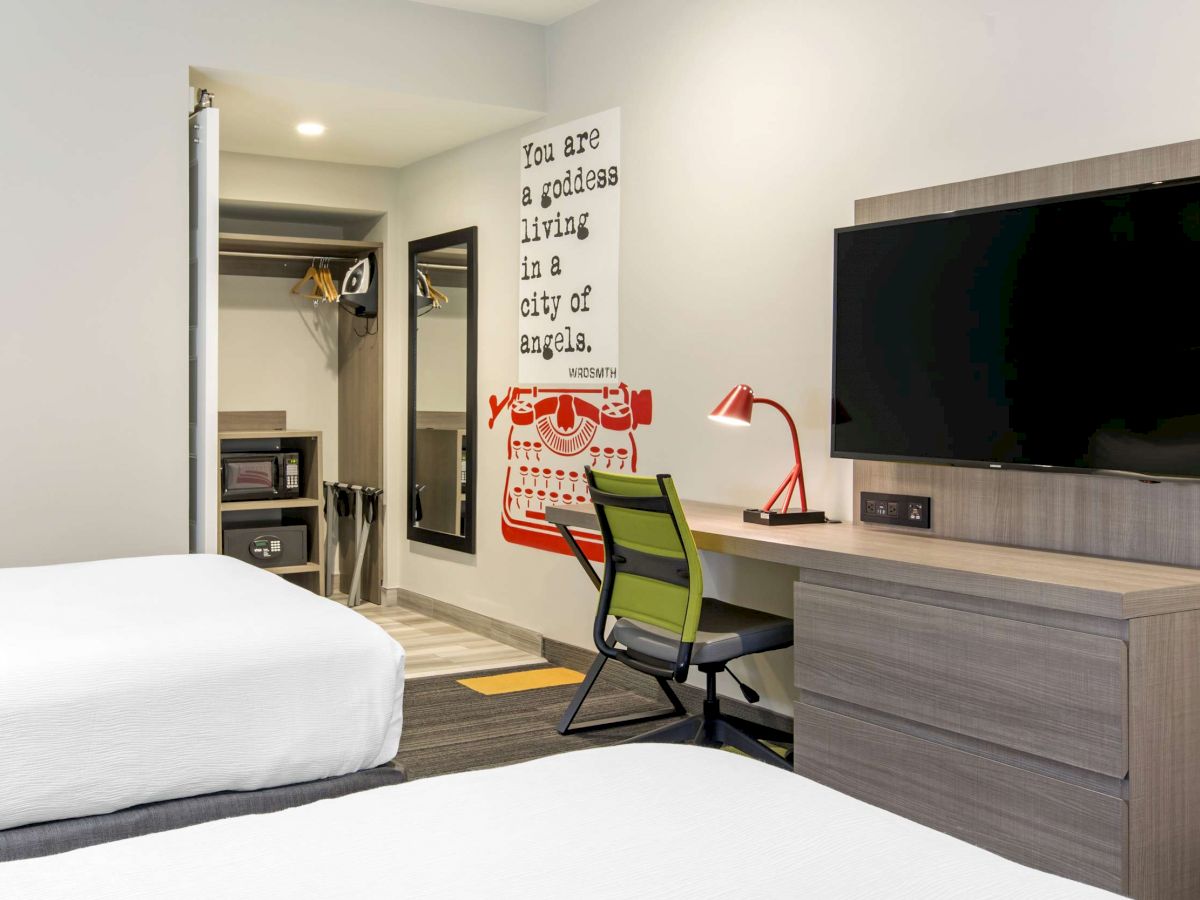 The image shows a modern hotel room with two beds, a wall-mounted TV, a desk with a red lamp, and an open closet area.