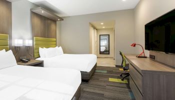 The image shows a modern hotel room with two double beds, a wall-mounted TV, a desk with a red lamp and chair, and a view towards the hallway.