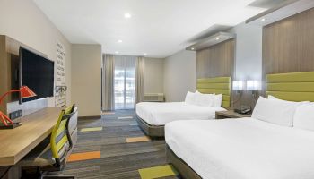 The image shows a modern hotel room with two queen beds, a desk with a red lamp, a chair, a wall-mounted TV, and patterned carpeting.