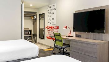 A modern hotel room with two beds, a wall-mounted TV, a desk with a chair and lamp, a closet, and a bold text wall decal.