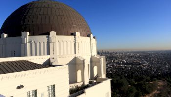 A white observatory with a large dome overlooks a sprawling cityscape under a clear blue sky, with skyscrapers visible in the distance.