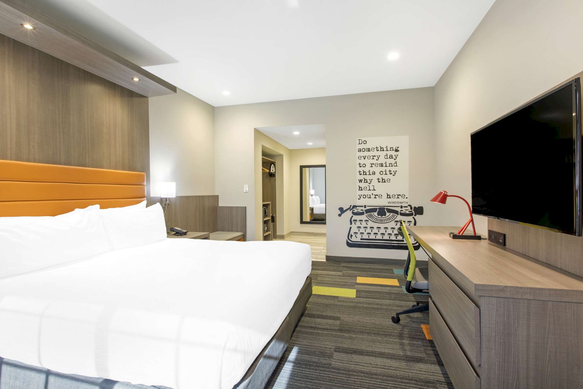 A modern hotel room with a large bed, desk, chair, TV, and wall art depicting a typewriter, with the saying, "Do something every day to remind this city why the heck you're here."