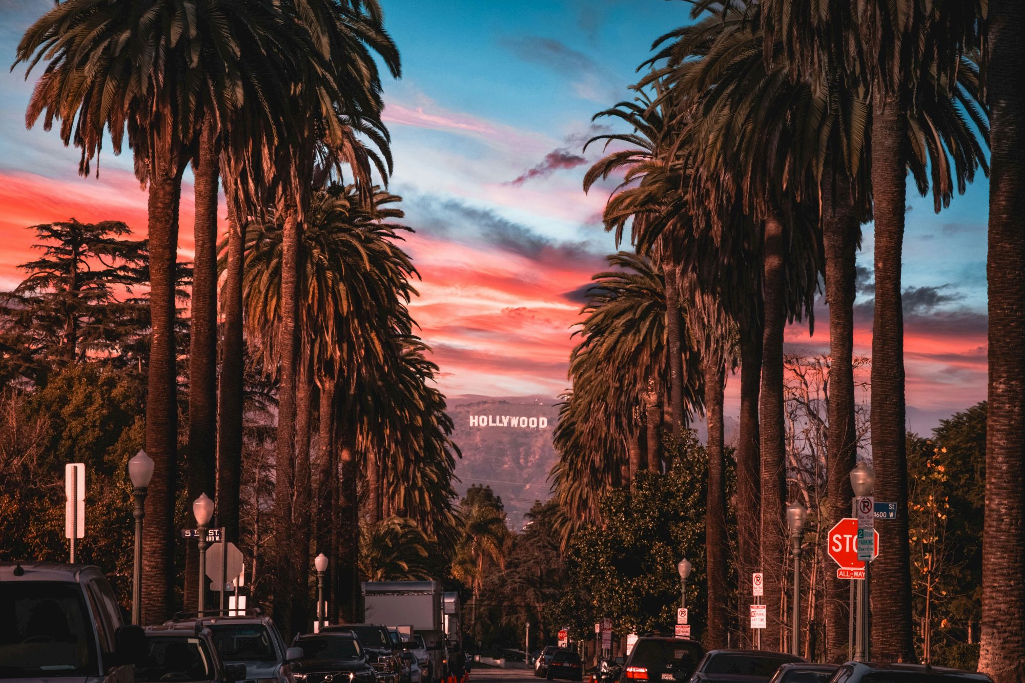 This image shows a street lined with tall palm trees during sunset, with the iconic Hollywood sign visible in the distance.