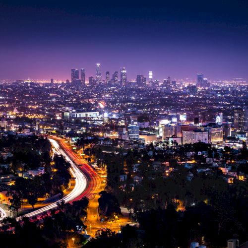 A nighttime cityscape features a brightly lit downtown skyline with tall buildings and a winding highway with busy traffic, surrounded by city lights.